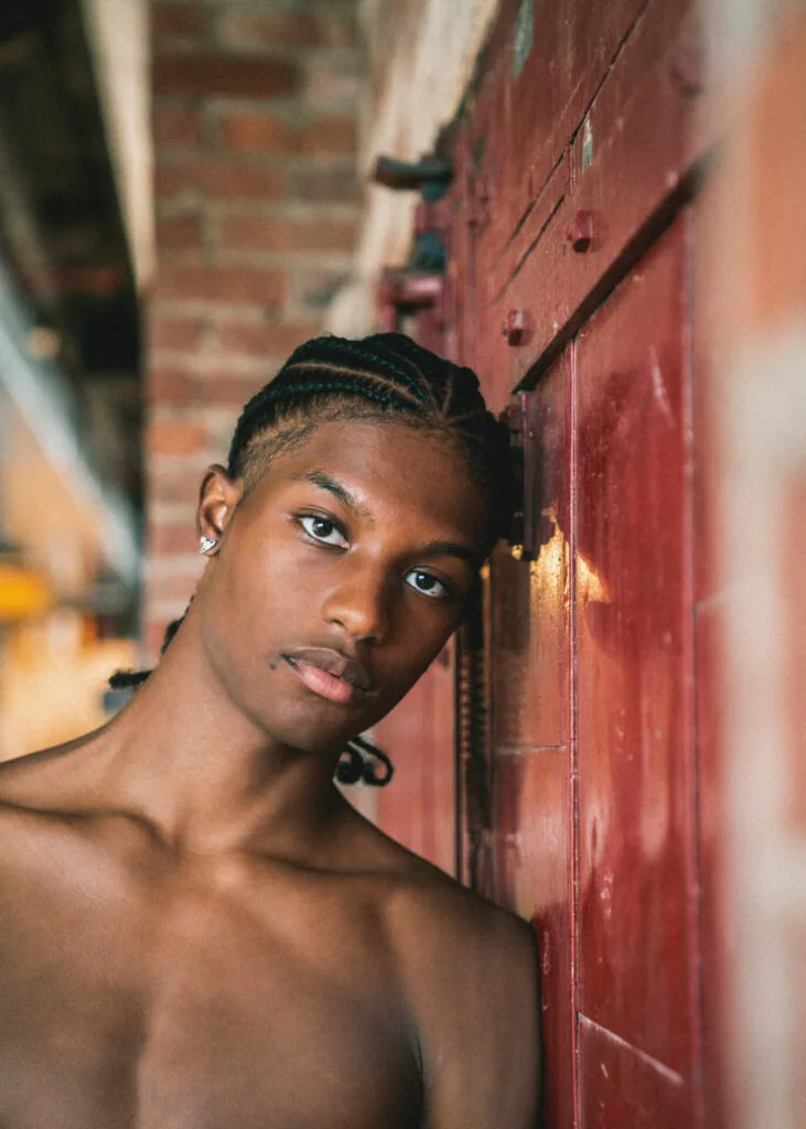 Male model with braided hair leaning against weathered red door in urban alley setting