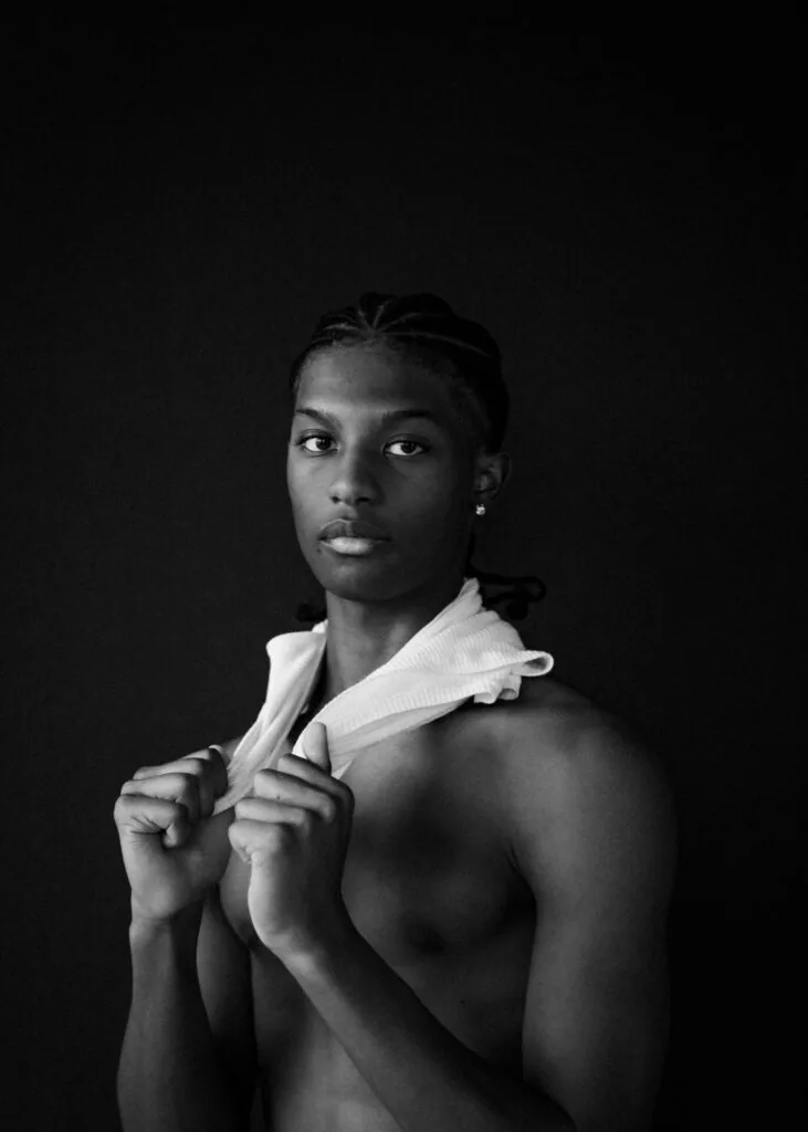 Male model shirtless against black background holding white draped fabric, direct gaze, professional portrait lighting