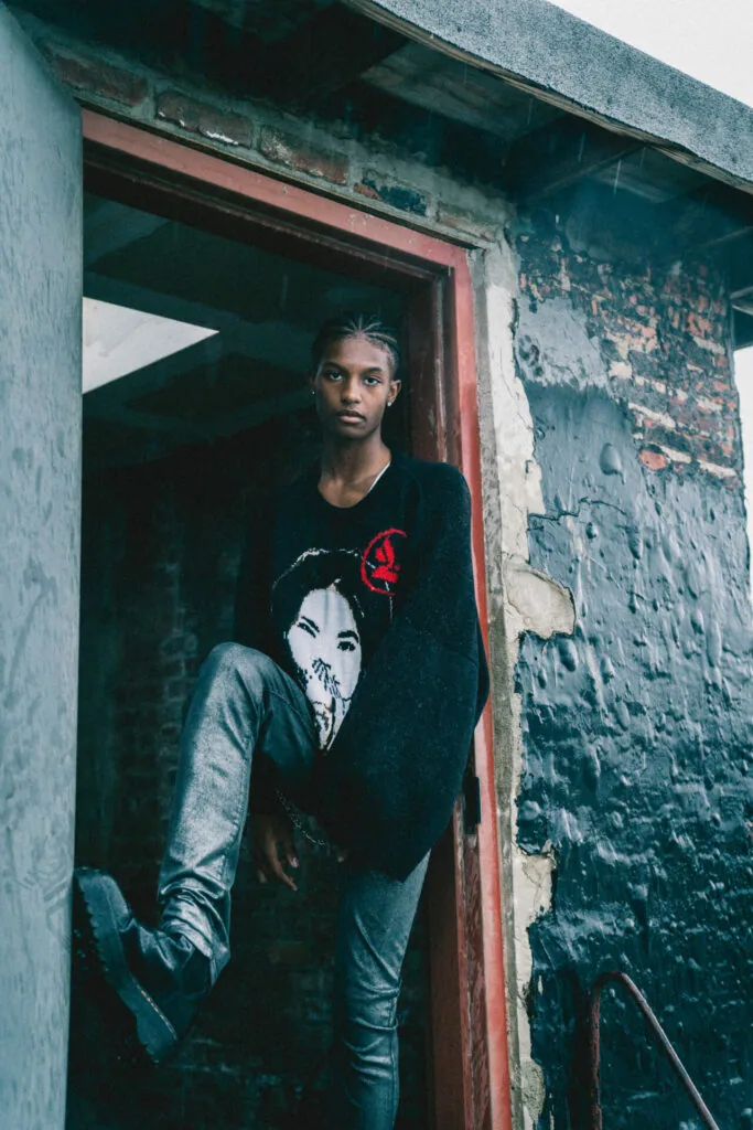 Male model in black graphic t-shirt standing in weathered doorway with red frame against peeling brick wall