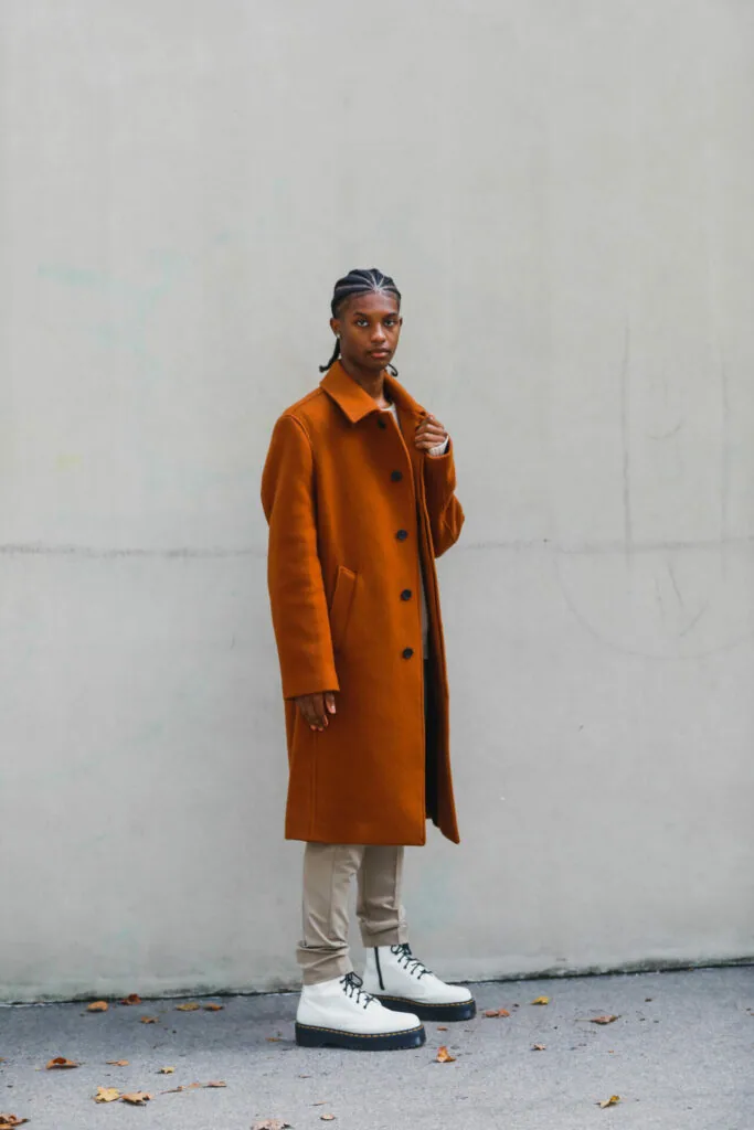 Person wearing rust-orange wool coat over cream trousers and white platform shoes against white wall
