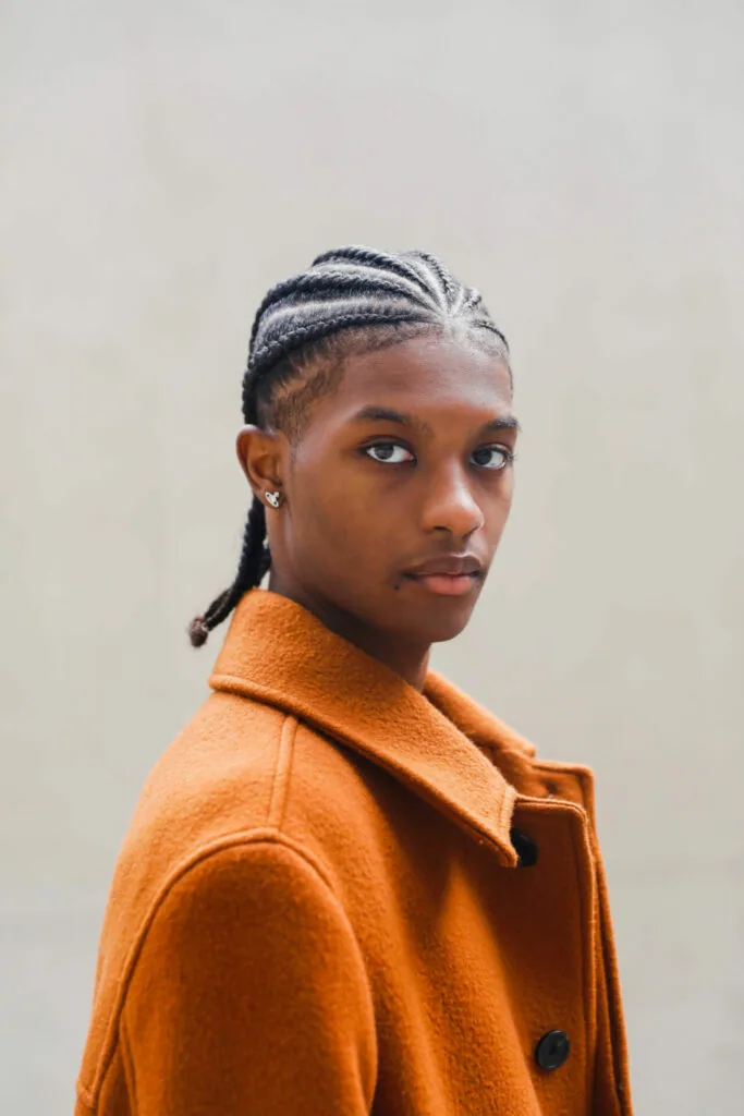 Young man wearing orange coat with braided hair and earring against neutral background