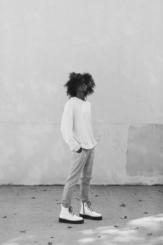 Person with curly dark hair wearing white long-sleeve shirt and light pants with platform lace-up boots standing against weathered white wall