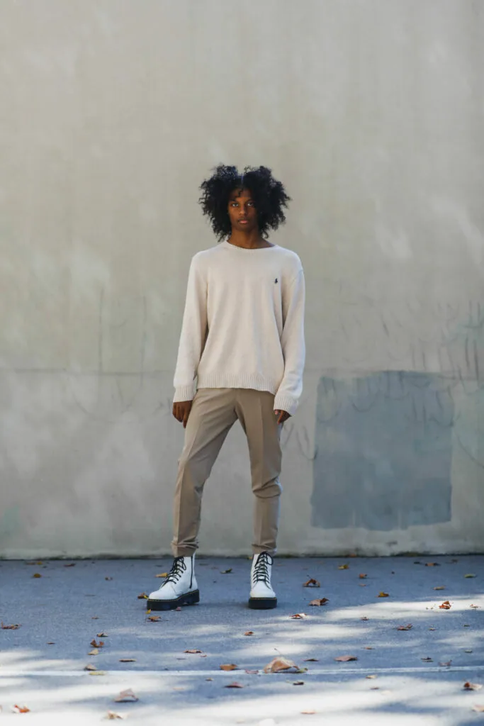 Male model with curly dark hair wearing cream sweater and khaki pants with white sneakers standing against white textured wall