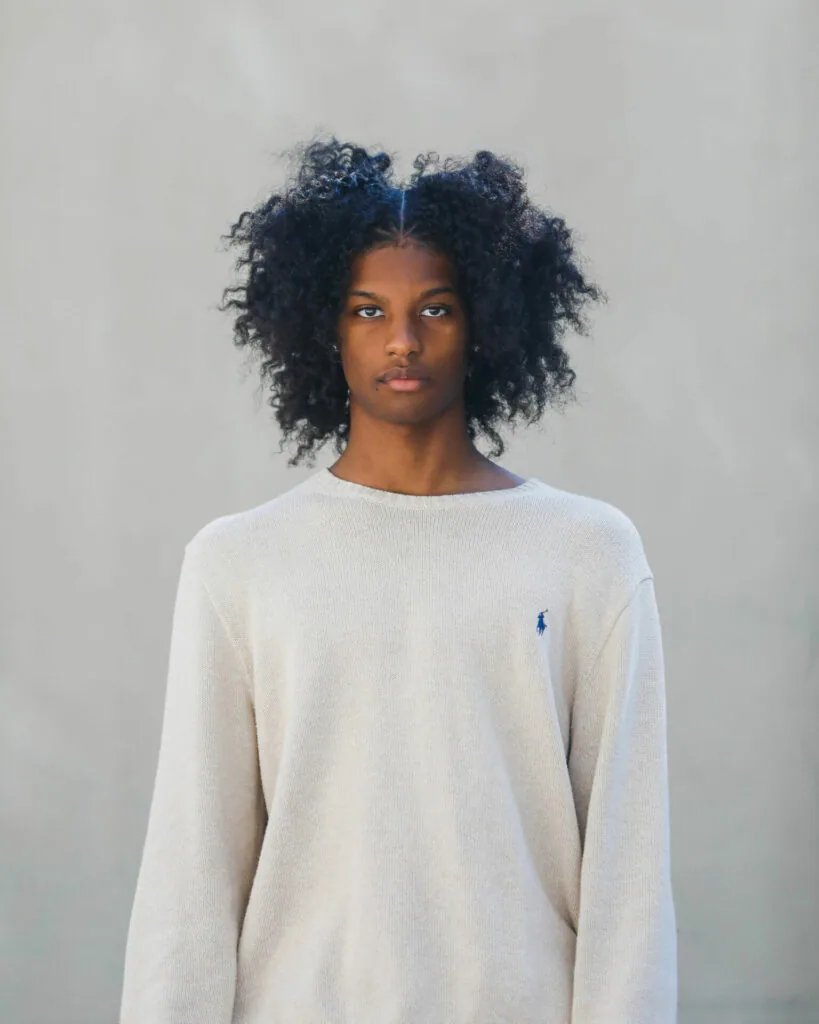 Person with voluminous dark curly hair wearing cream knit crewneck sweater against neutral background