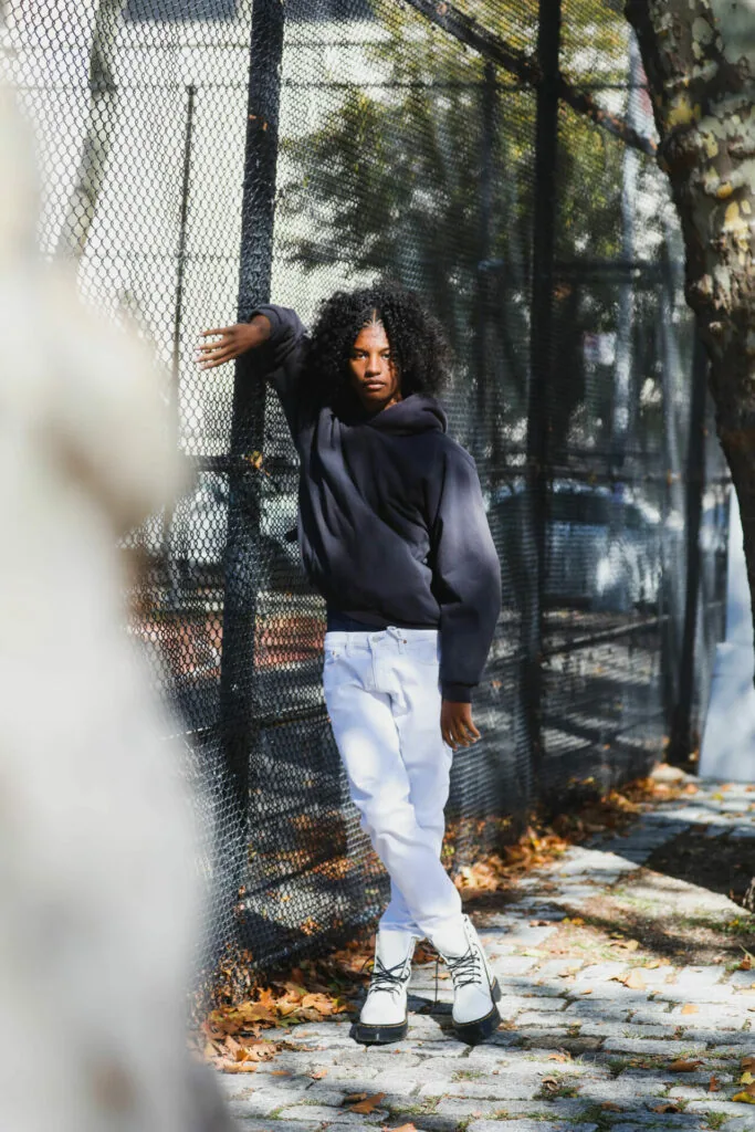 Woman in navy hoodie and white jeans standing against chain-link fence in sunlit outdoor setting with autumn leaves