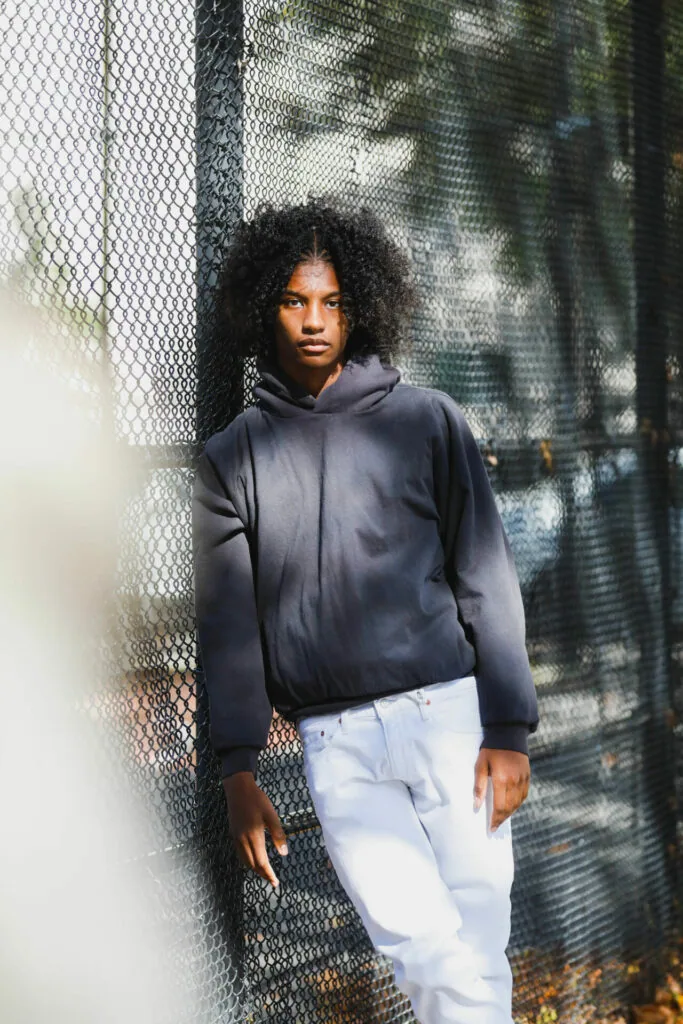 Male model wearing black hoodie and white pants posing against chain-link fence with dappled shadow backdrop