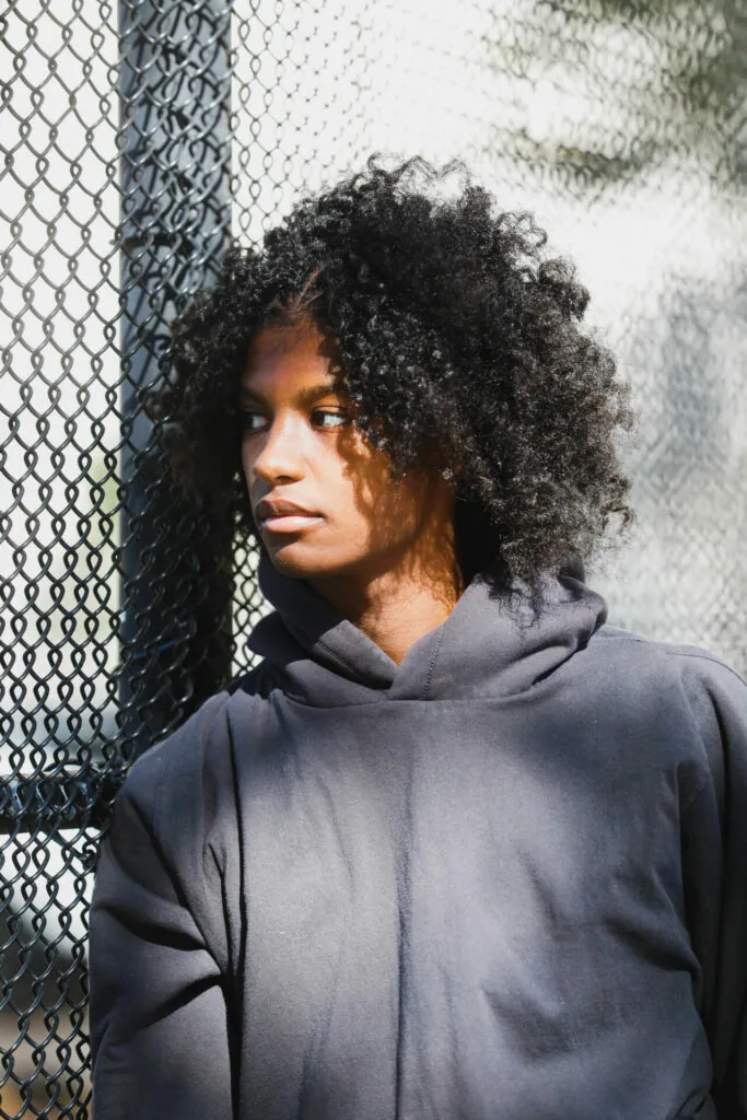 Person with textured curly hair wearing dark hoodie standing against chain-link fence in urban setting