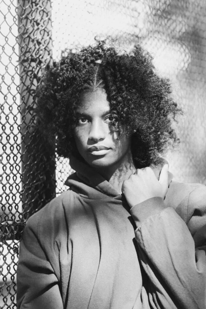 Man with voluminous curly hair wearing collared shirt against chain-link fence in black and white portrait