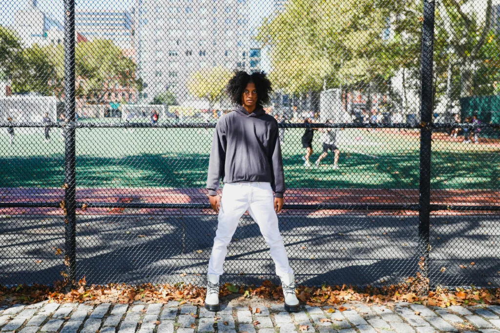 Male model in gray hoodie and white joggers standing at tennis court fence with fall leaves on ground