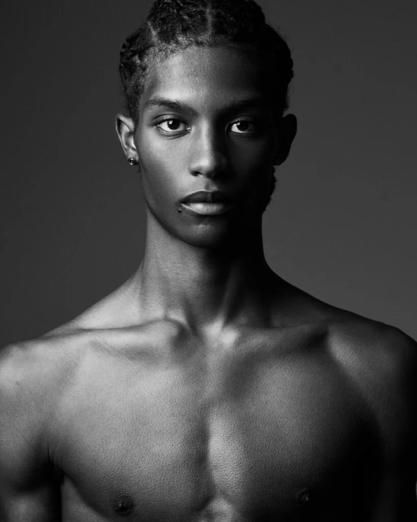 Male model shirtless with defined chest, short styled hair, and ear piercing against neutral gray background in professional fashion photography