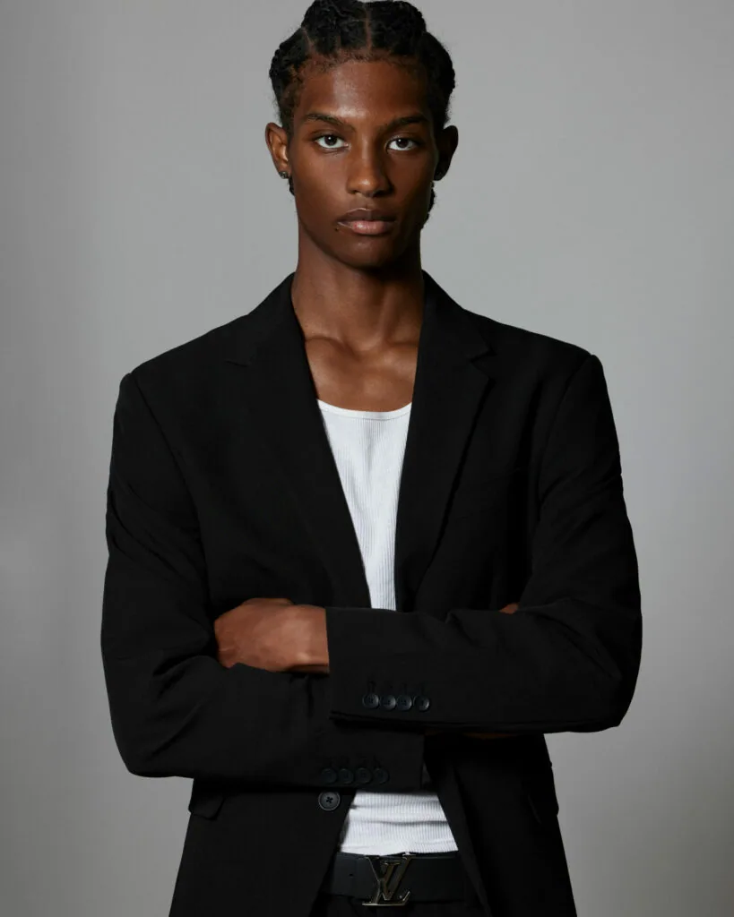 Male model in black blazer and white ribbed shirt, arms crossed, professional studio portrait with neutral gray background