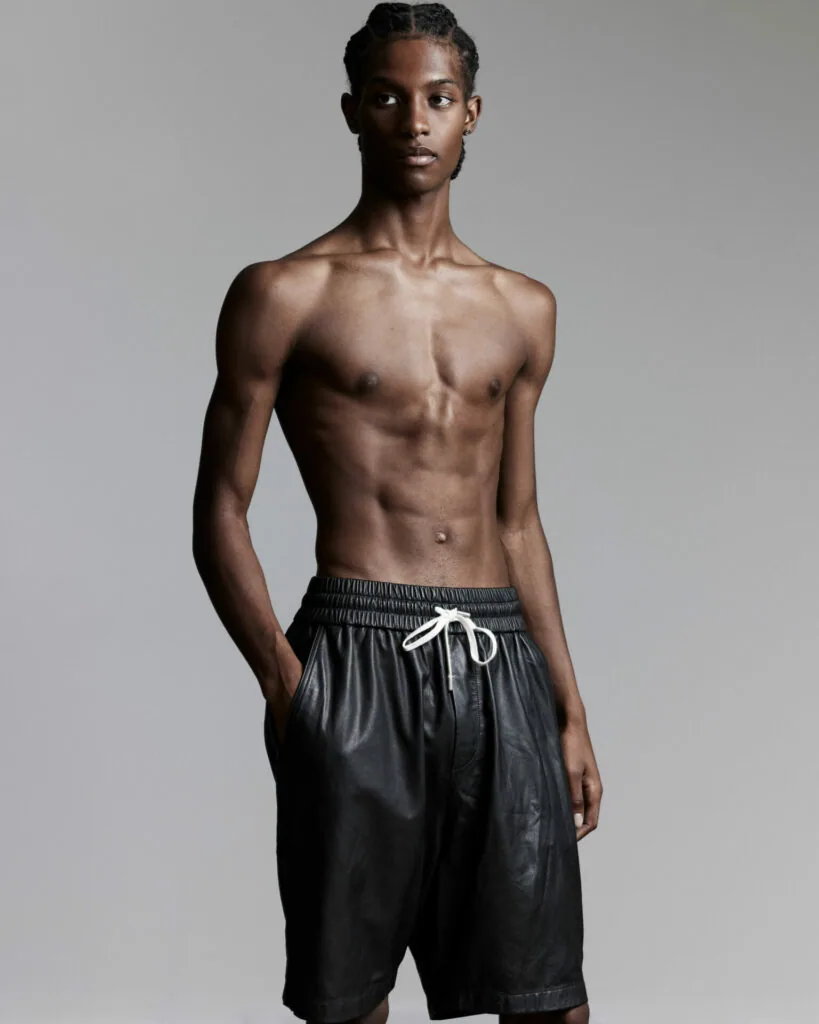Shirtless male model wearing black drawstring shorts against gray background in professional fashion photography