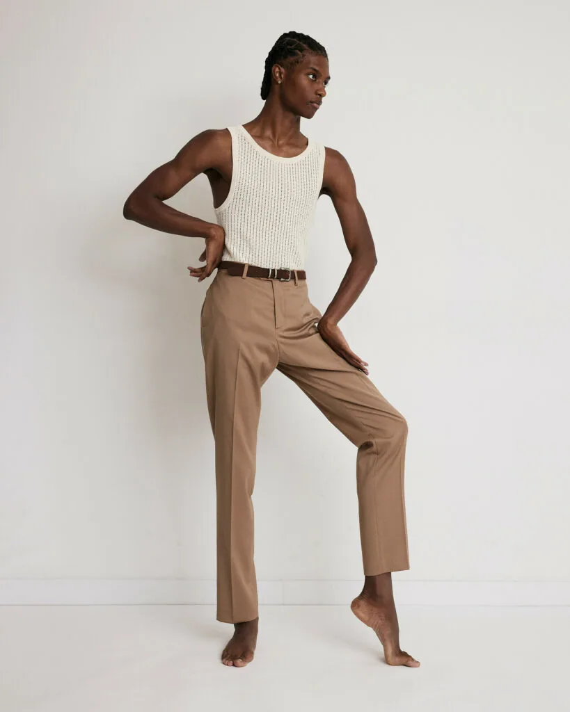 Male model in cream ribbed tank top and tan trousers with brown belt, posing barefoot against white background.