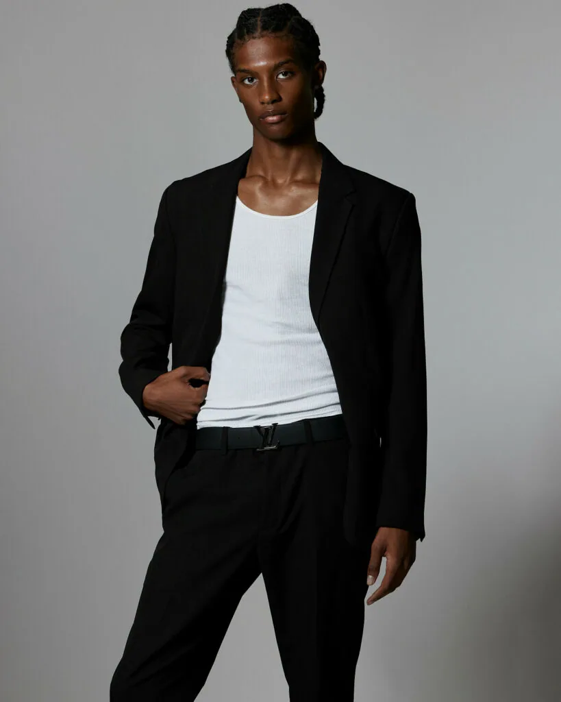 Male model wearing black blazer, white ribbed tank top, and black trousers against neutral background