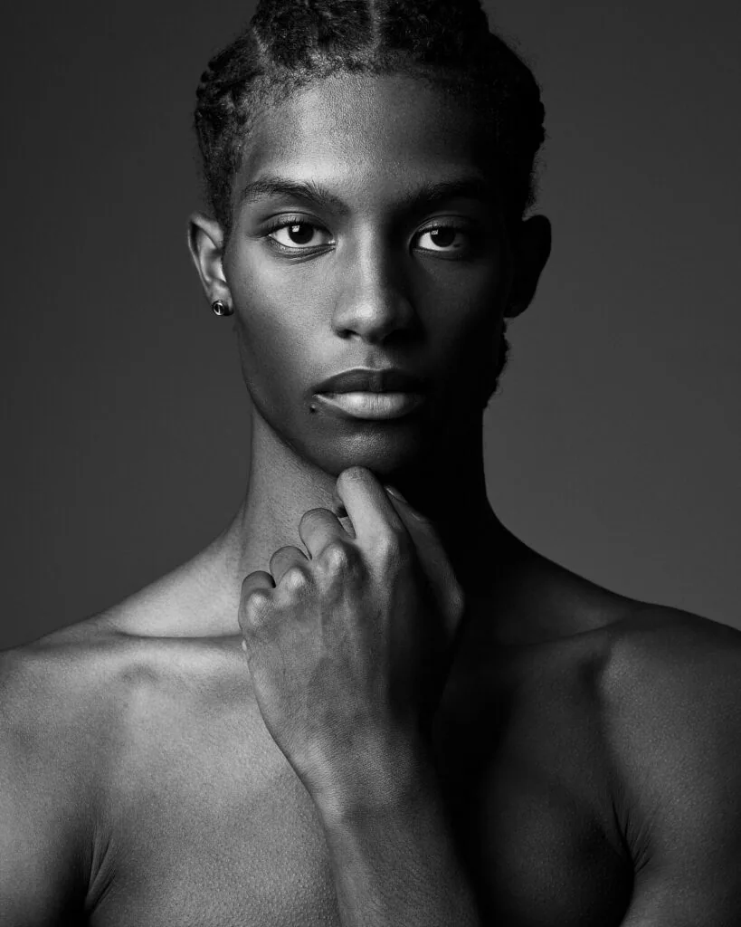 Male model with short dark hair wearing ear stud, hand to chin, against gray background in black and white portrait