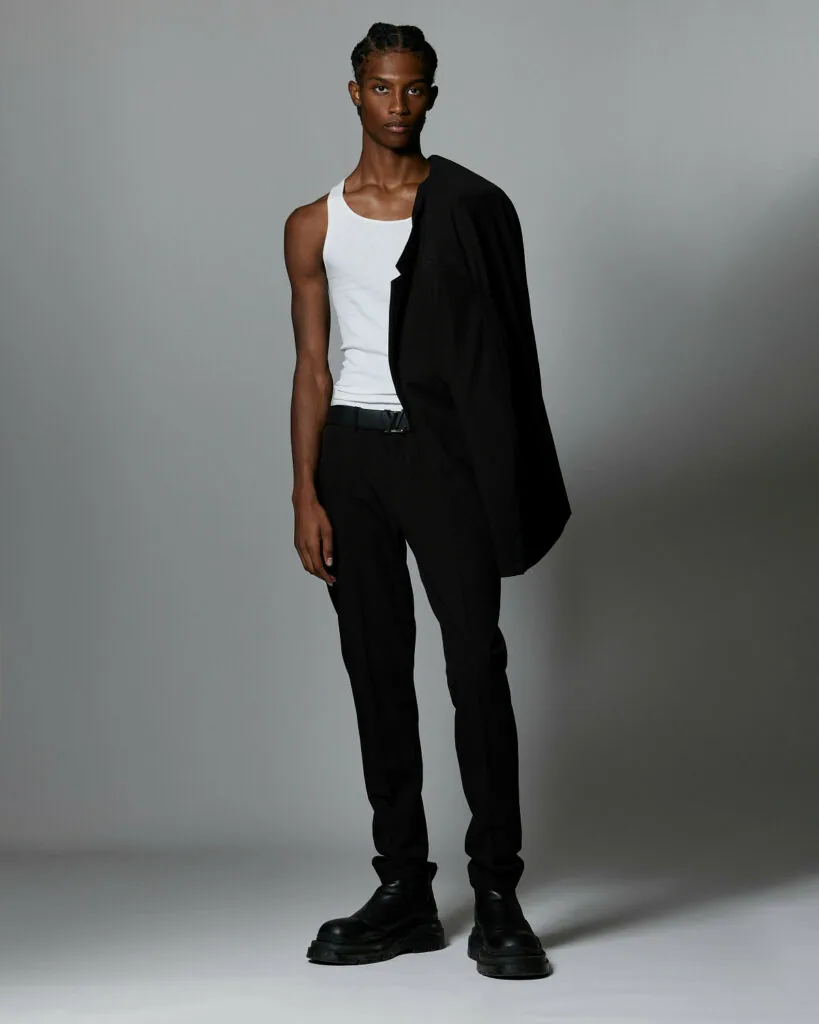 Male model wearing white sleeveless tank top, black cardigan, and black pants with black boots against gray backdrop.