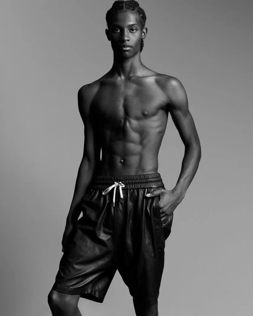 Shirtless male model in black drawstring shorts against gray background, professional fashion portrait
