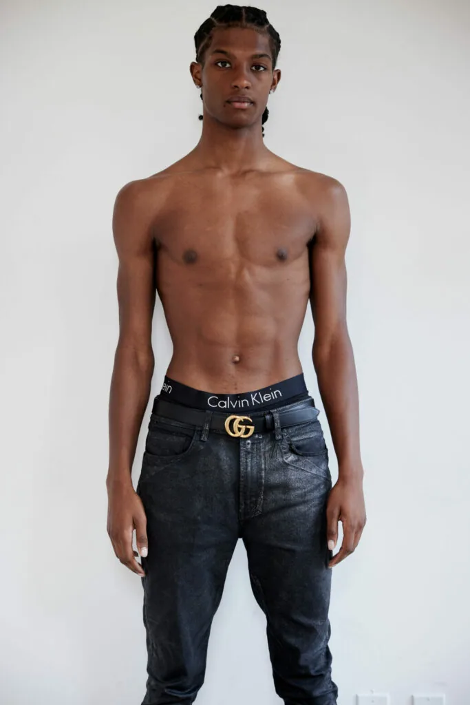 Shirtless male model wearing black jeans with branded waistband and gold belt buckle against white background
