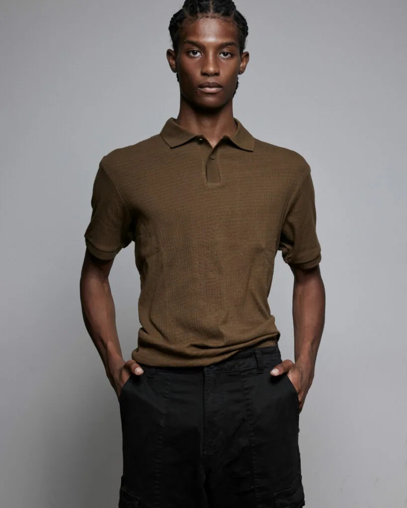 Male model wearing olive brown polo shirt and black trousers against gray background