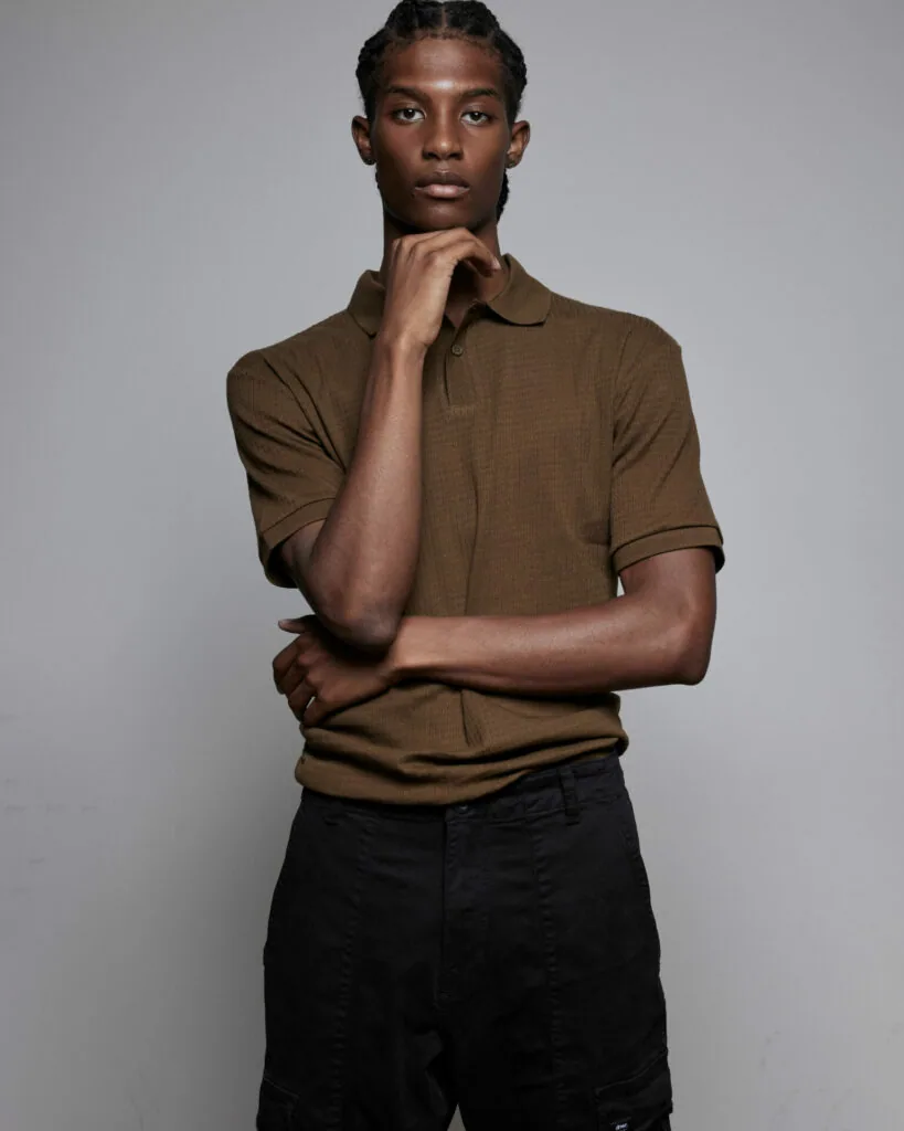 Male model in brown polo shirt and black pants posing against gray background