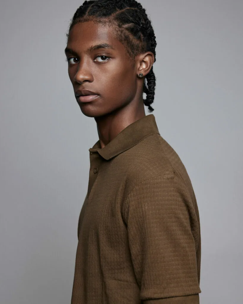 Young male model with braided hair wearing brown textured polo shirt against neutral gray background
