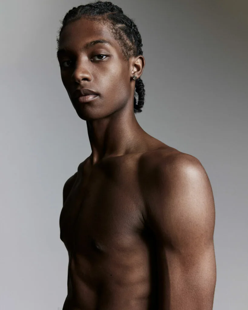 Shirtless male model with braided hair in professional studio portrait against neutral gray background