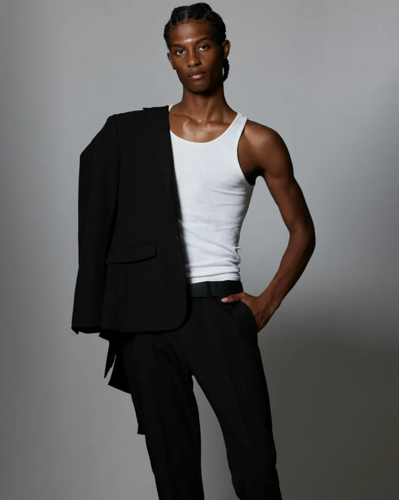 Male model wearing white tank top and black blazer with black pants against gray background