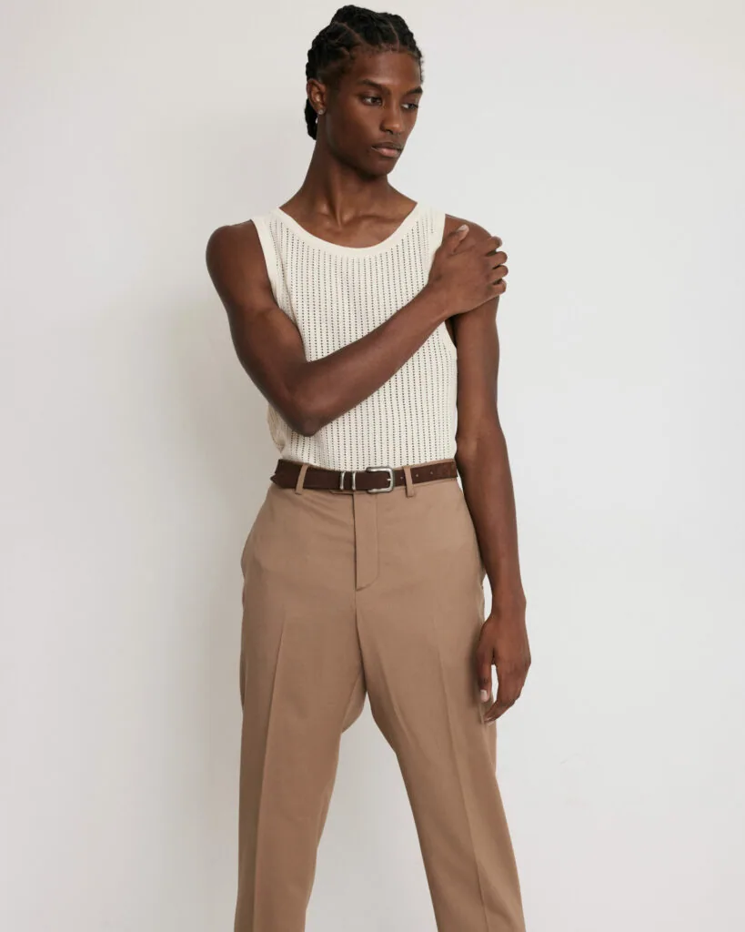 Male model in cream ribbed tank top and tan trousers with brown belt against white background