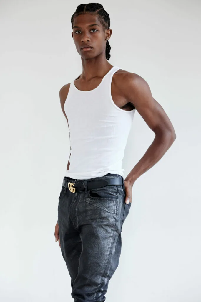 Male model in white sleeveless tank top and dark jeans with gold belt buckle against white background
