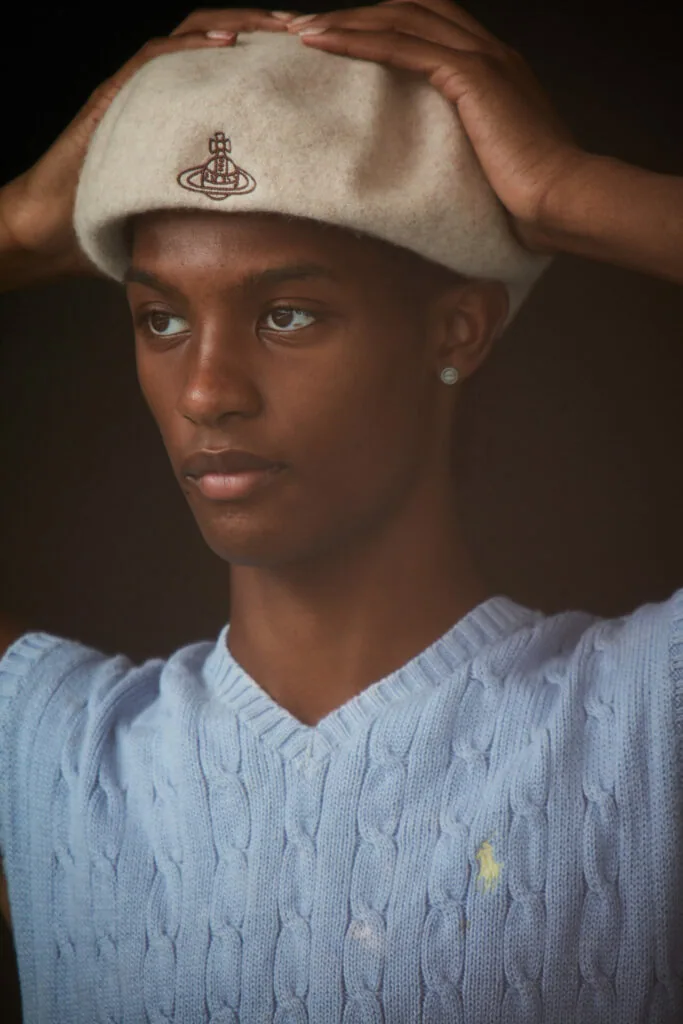 Male model wearing cream beret with embroidered logo and light blue cable-knit sweater, hands adjusting hat against dark background.