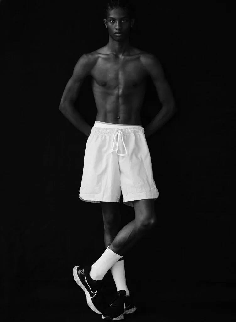 Shirtless male model in white athletic shorts and white socks with black sneakers against black background