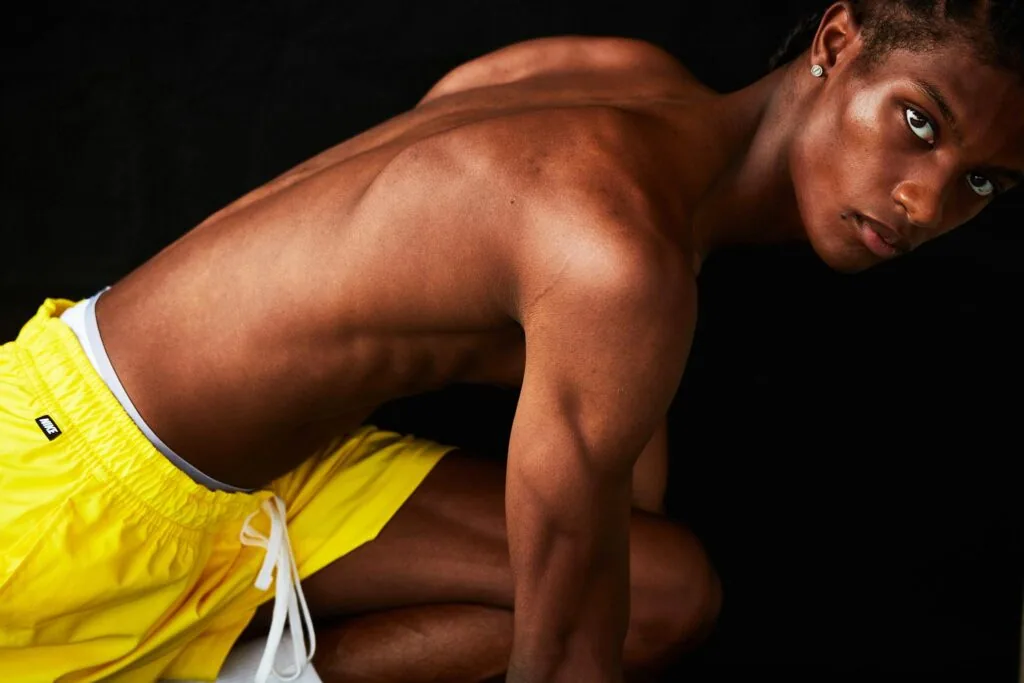 Shirtless male model in yellow athletic shorts against black background, showcasing muscular physique in professional studio lighting