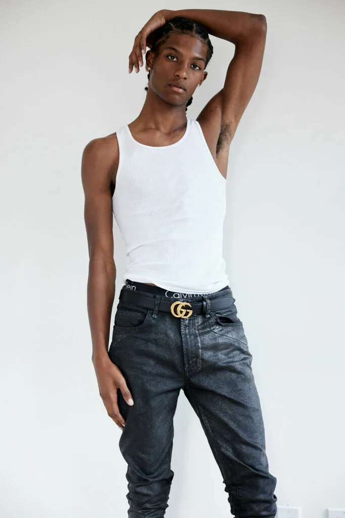 Male model wearing white sleeveless tank top and dark jeans with gold buckle belt against white background