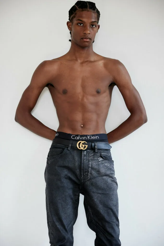 Shirtless male model wearing dark denim jeans with designer belt against white studio backdrop