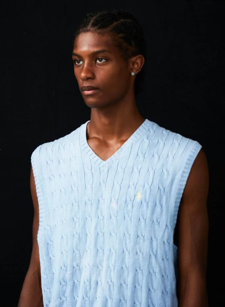 Young male model wearing light blue cable-knit sleeveless vest against black background studio portrait