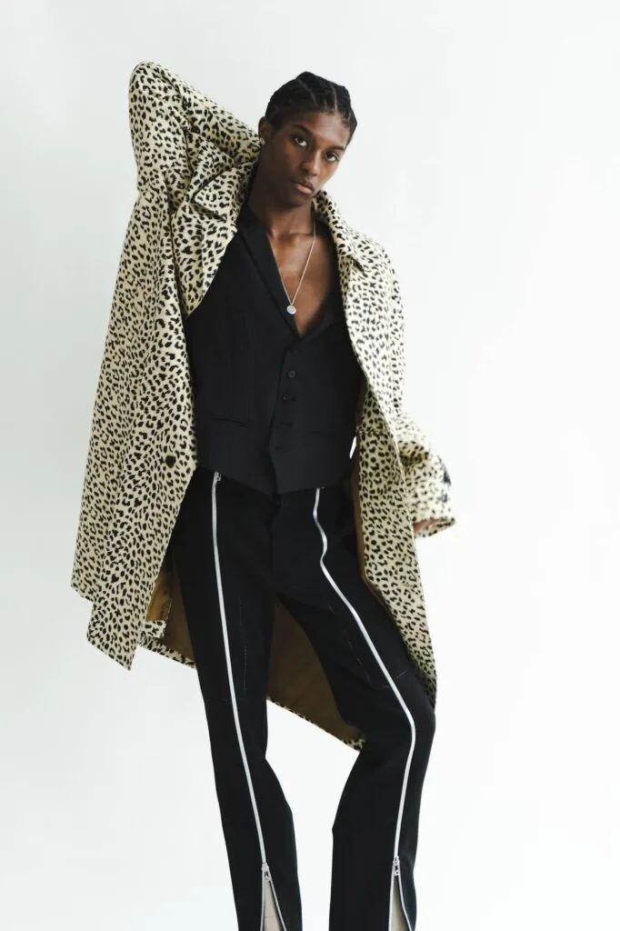 Male model wearing leopard print cardigan over black blazer and black pants with side stripes, posed against white background