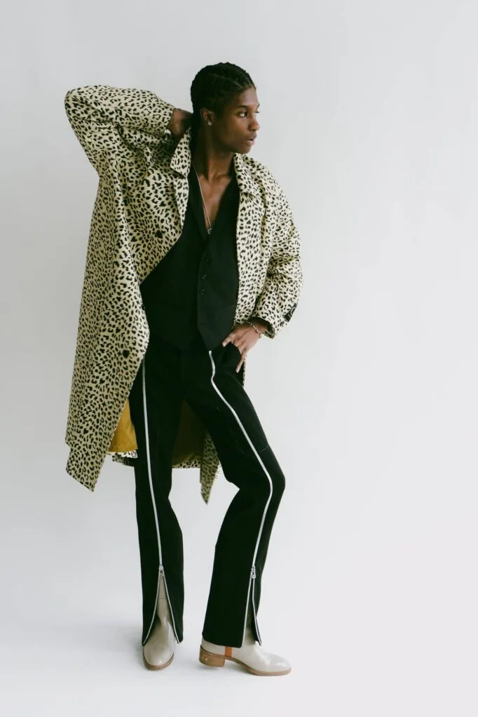 Male model in leopard print coat and black zippered pants with white sneakers poses against white background