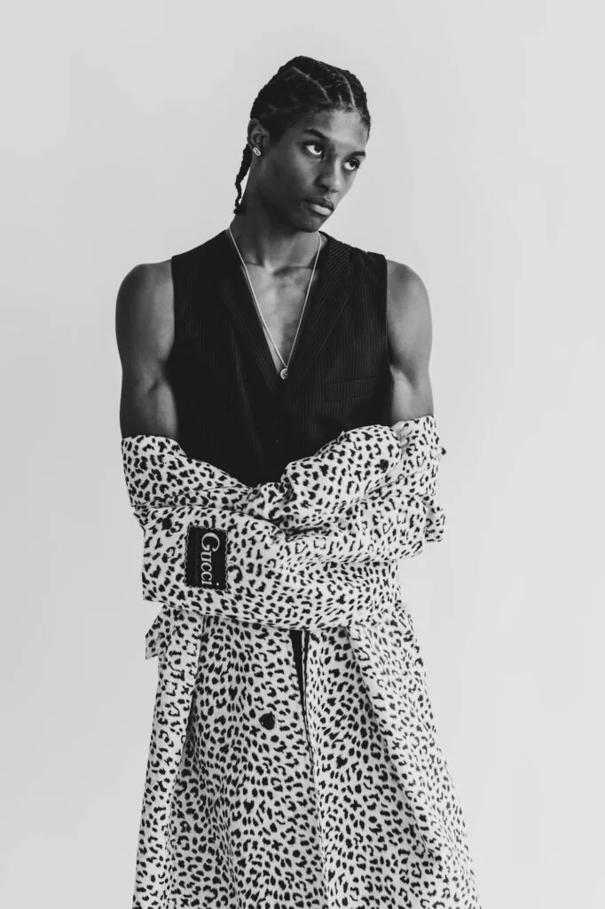 Male model wearing black ribbed vest over white spotted wrap skirt, studio portrait against neutral background