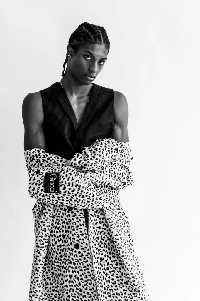 Male model wearing black sleeveless vest and leopard-print skirt against white background in professional fashion portrait