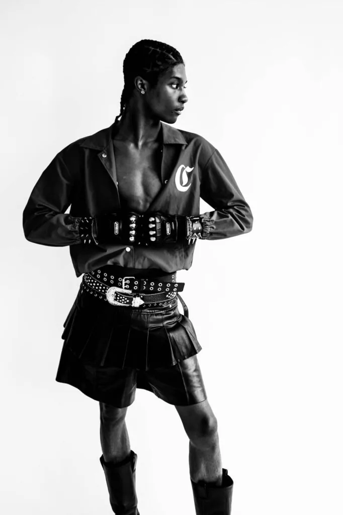 Male model in black jacket with studs, leather shorts, and studded gloves against white background