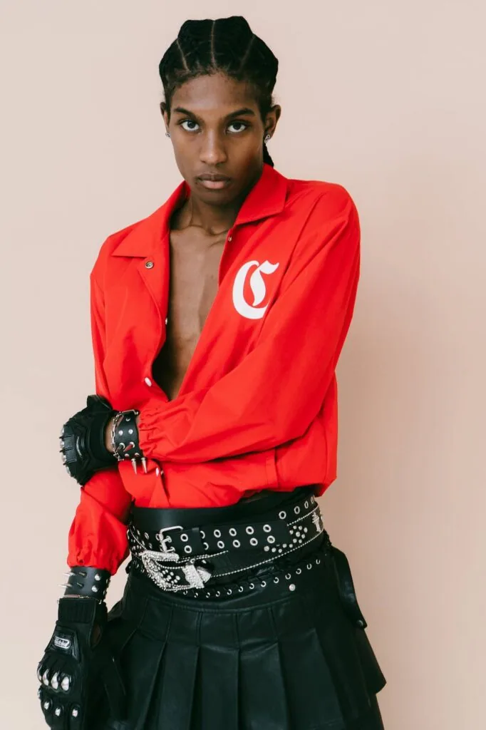 Male model in red windbreaker with white logo, black studded belt, and pleated black skirt with leather gloves against neutral background.