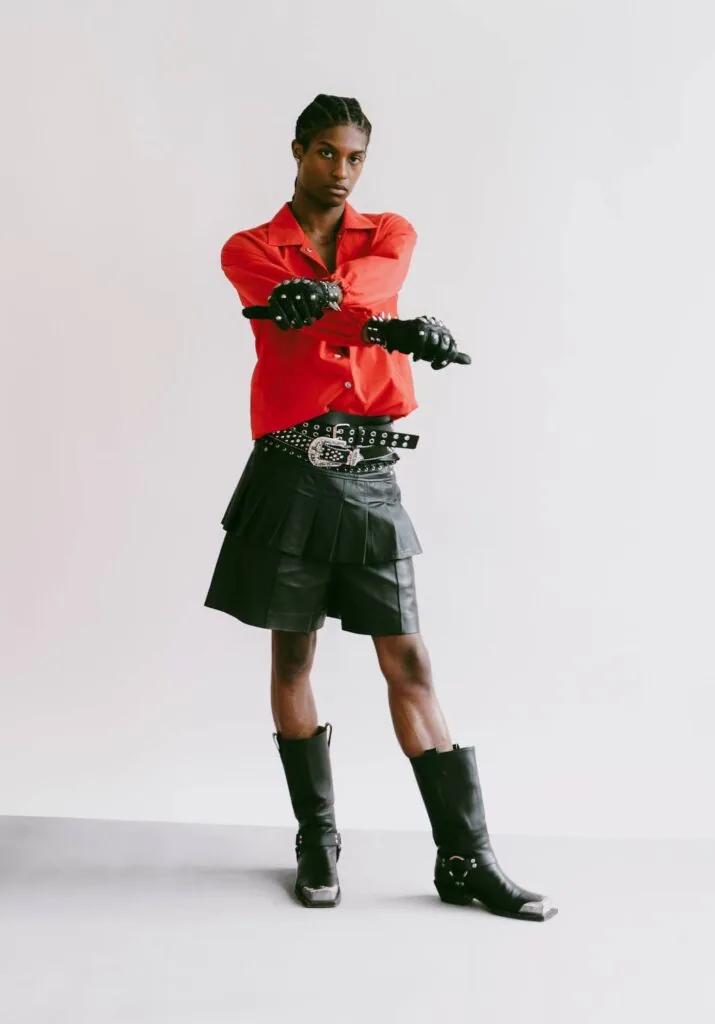 Male model in red satin shirt, black studded belt, and black cargo shorts with matching boots against white background
