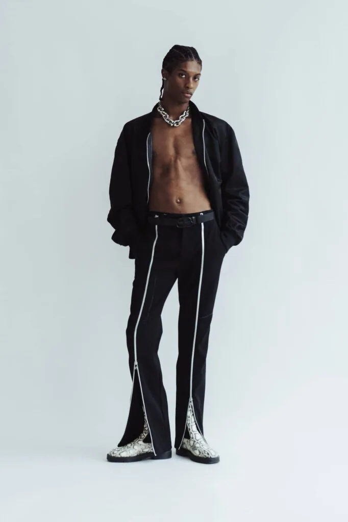 Male model wearing open black jacket, chain necklace, black pants with white side stripes and metallic boots against white background