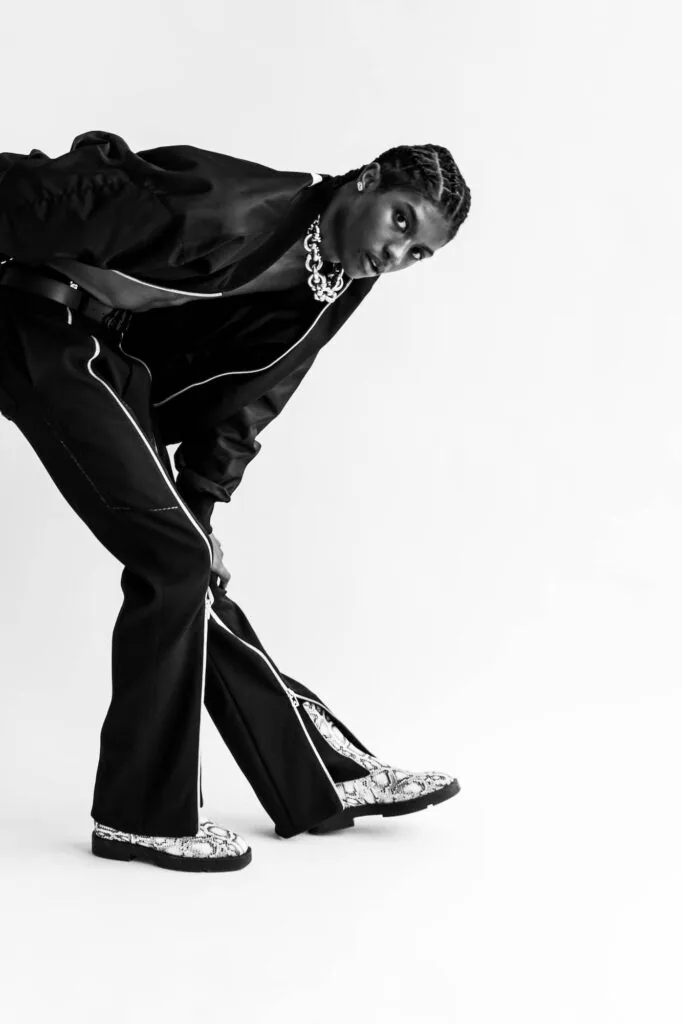 Male model in black oversized jacket and flared pants with metallic loafers, posed dynamically against white background