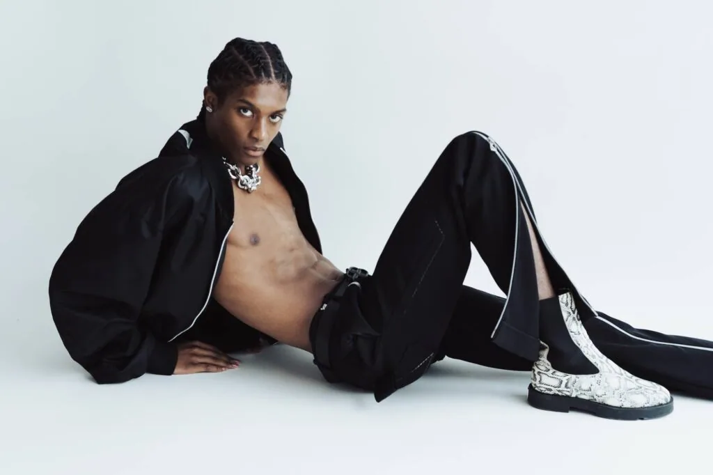Male model in black blazer and track pants with silver chain necklace, seated pose on white background