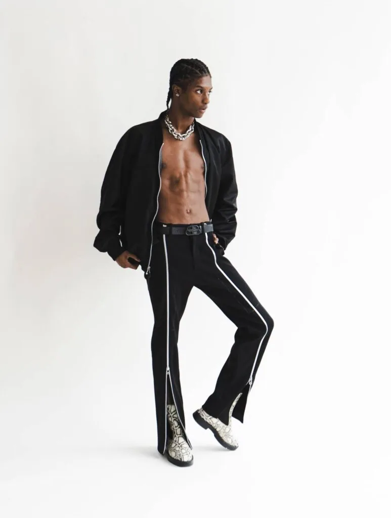 Male model in black zip jacket and piped pants with chain necklace against white background