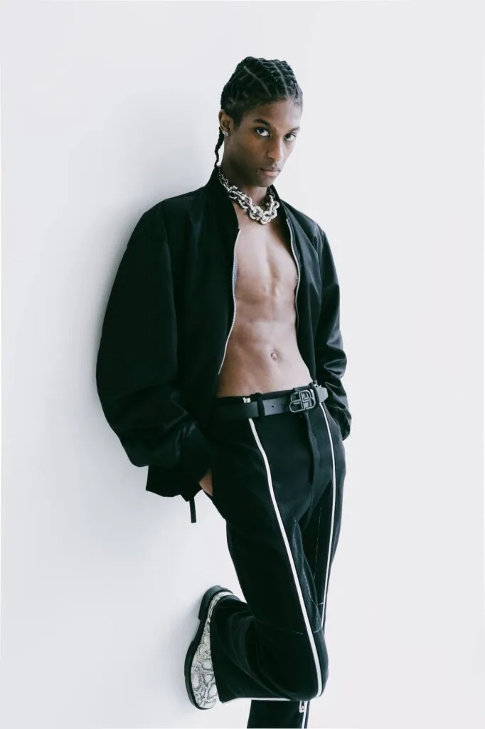 Male model wearing black open shirt and black cargo pants with silver chain necklace against white background