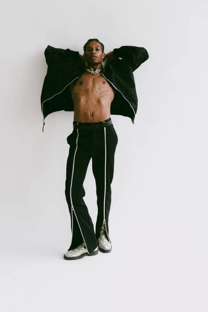 Male model shirtless wearing open black jacket with silver chain necklace and black pants with piping against white background