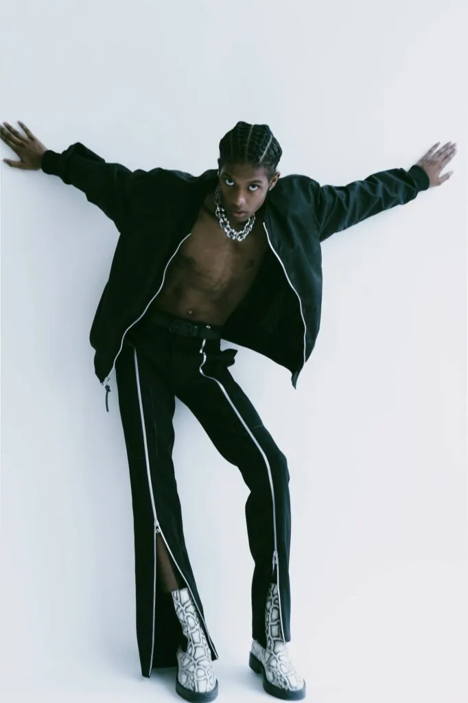 Male model in black jacket and piped pants with arms outstretched, wearing chain necklace and patterned boots against white backdrop