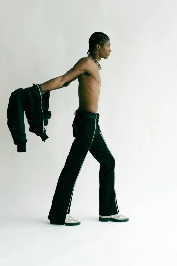 Shirtless male model in black pants with white trim and sneakers holding black jacket, profile view against white background.
